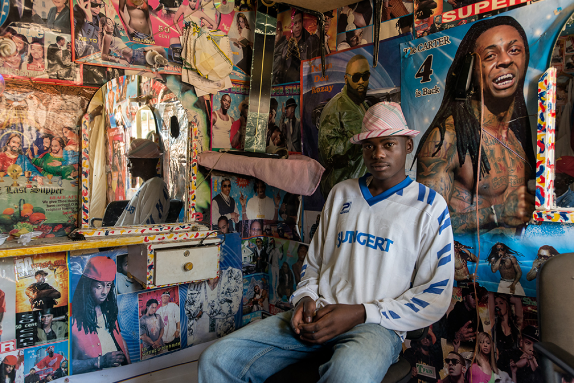 A young man sits inside a hair salon whose walls are covered in posters of Black musicians and other pop culture figures.