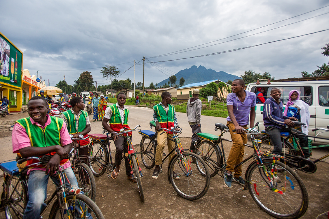 Young men wearing green vests pause on their bicycle taxis at the side of a road.