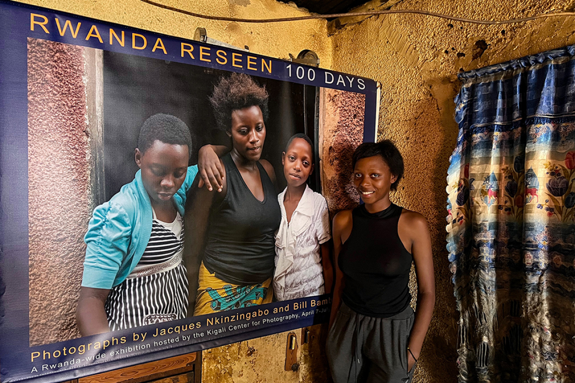 A young woman stands inside her home next to a large printed photograph from the Rwanda Reseen exhibition.