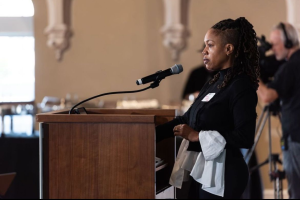 A woman stands at a wooden podium speaking into a microphone in a large, bright room.