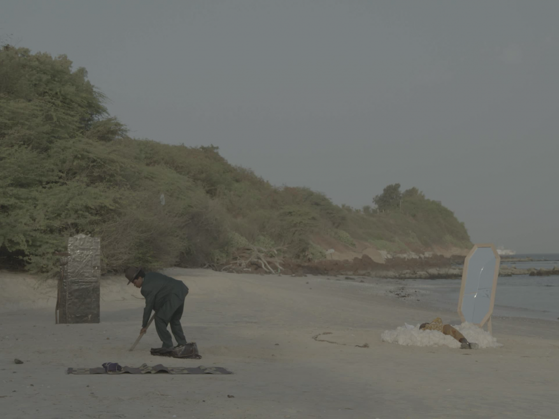 A person on the left digs into sand on a beach, with another person on the right lying next to a broken mirror next to water