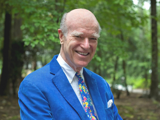 Joel Fleishman poses for a photo outdoors, with woods in the background; he wears a bright blue blazer, white shirt and colorful printed tie.