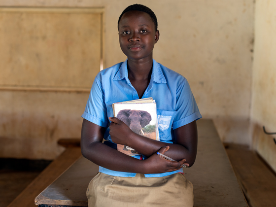 A young woman in a classroom holds a notebook and pen.